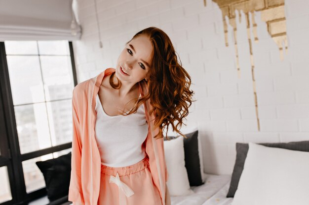 Dame intéressée aux cheveux ondulés exprimant des émotions positives. Jocund fille blanche en pyjama souriant.