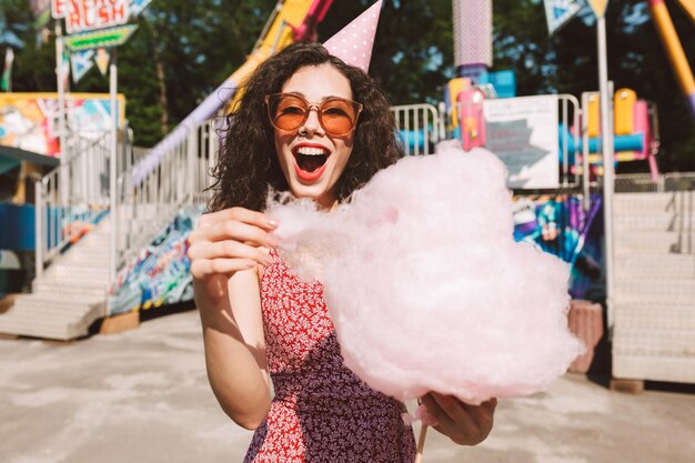 Dame joyeuse avec des cheveux bouclés noirs dans des lunettes de soleil et une casquette d'anniversaire debout avec de la barbe à papa à la main et regardant joyeusement à huis clos tout en passant du temps dans un parc d'attractions