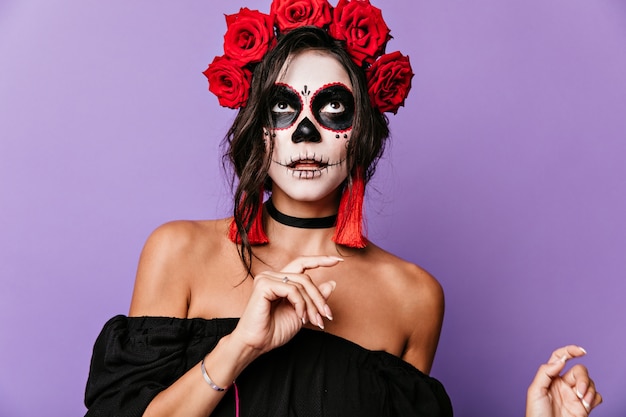 Photo gratuite dame latine bronzée en pensée sur un mur lilas. fille aux cheveux noirs bouclés et roses en eux posant avec un masque squelette sur son visage