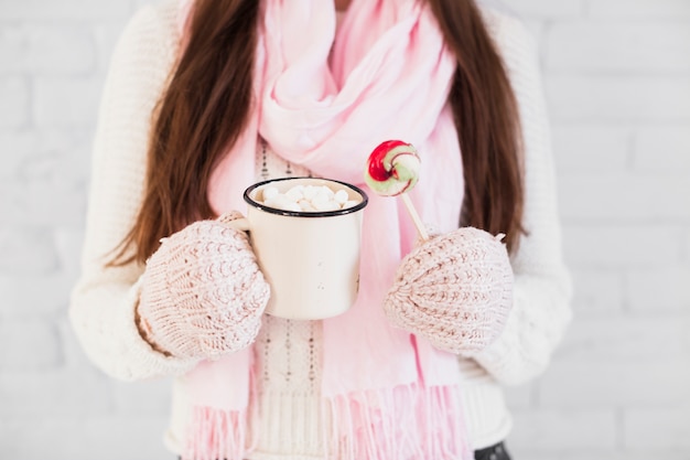 Dame en mitaines et foulard tenant une tasse avec des guimauves et sucette