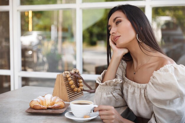 Dame à la mode dans un café, boire du café, s'appuyer sur la table et profiter de la vue à l'extérieur.