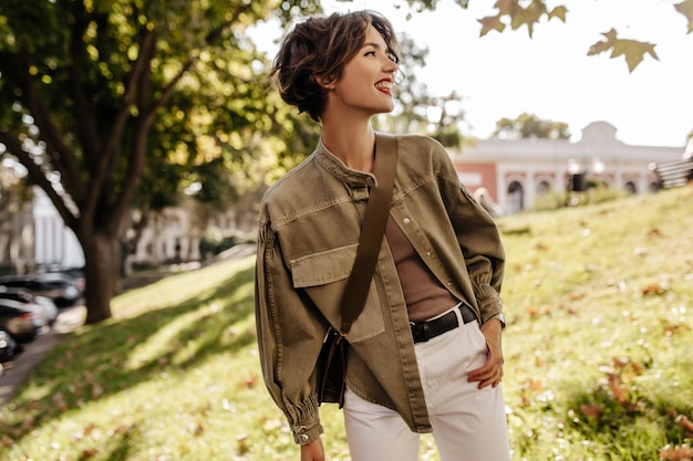 Dame moderne en veste en jean avec sac à main à l'extérieur. Femme aux cheveux ondulés avec des lèvres rouges en pantalon blanc souriant à l'extérieur.