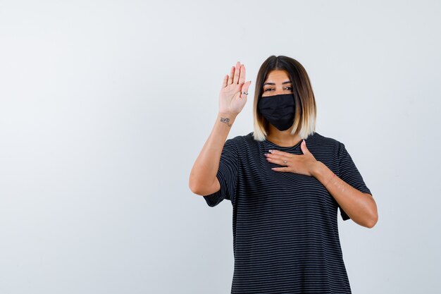 Dame montrant le geste d'arrêt en robe noire, masque médical et à la politesse. vue de face.