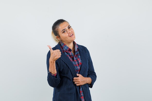 Dame montrant le pouce vers le haut en chemise, veste et à la vue confiante, de face.