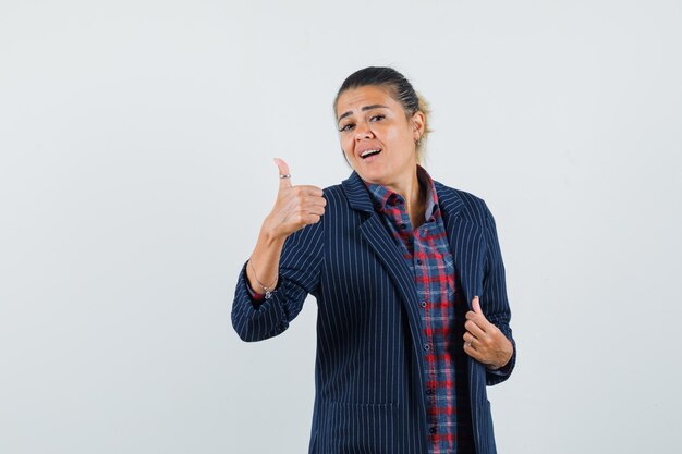 Dame montrant le pouce vers le haut en chemise, veste et à la vue confiante, de face.