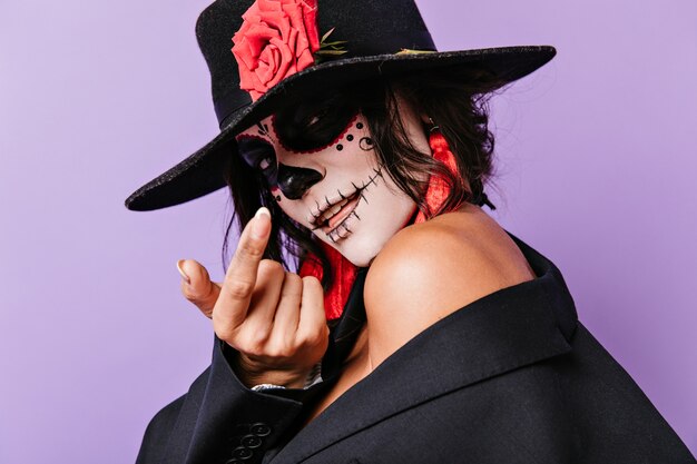 Une dame mystérieuse avec une rose sur un chapeau fait signe au doigt de l'approcher. Closeup portrait de jolie brune avec des boucles d'oreilles rouges.