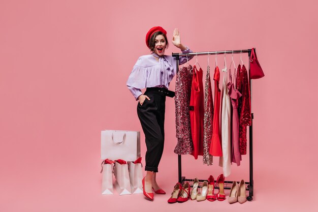 Dame pose dans le vestiaire avec des vêtements et des chaussures lumineux. Fille en béret et chemisier lilas regardant la caméra sur fond rose.