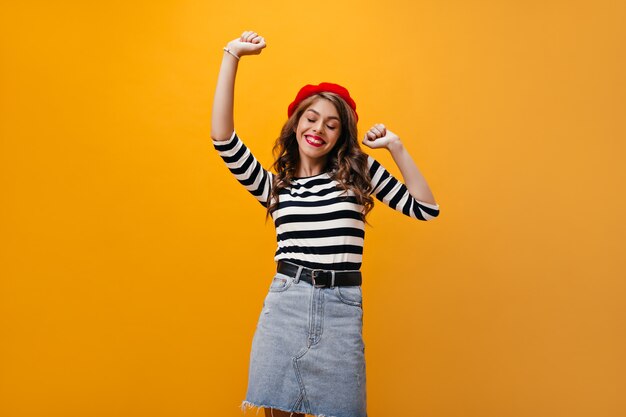 Dame positive en béret et chemise souriant sur fond orange. Femme élégante de bonne humeur avec des cheveux ondulés dansant sur fond isolé.