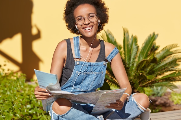 Photo gratuite dame positive à la peau foncée avec une coiffure afro, tient une carte, aime les voyages en vacances, veut atteindre certaines destinations, porte des combinaisons décontractées, des modèles en plein air dans un cadre tropical. les gens et les voyages