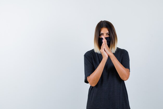 Dame pressant les mains ensemble tout en priant en robe noire, masque médical et à la recherche d'espoir. vue de face.