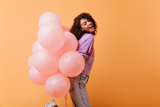 Dame rêveuse en tenue décontractée dansant avec un tas de ballons à l'hélium. Fille noire de bonne humeur se prépare pour la fête d'anniversaire.