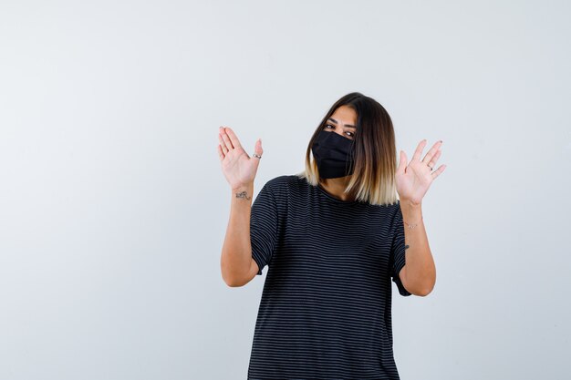 Dame en robe noire, masque médical montrant le geste d'arrêt et regardant attentivement, vue de face.