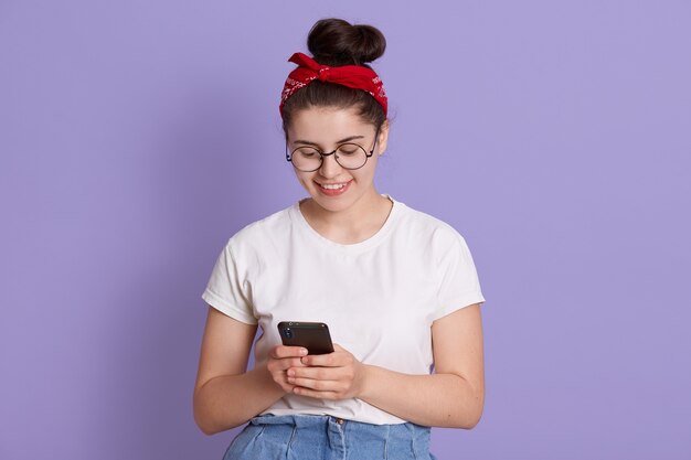 Dame souriante aux cheveux noirs et noeud tenant un téléphone intelligent dans les mains et lisant le message du petit ami, a l'air heureux, vêtu d'une tenue décontractée