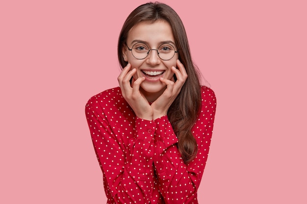 Dame souriante positive avec un regard doux, garde les mains sur les joues, porte des lunettes rondes, une blouse à la mode rouge, heureuse de faire un nouvel achat