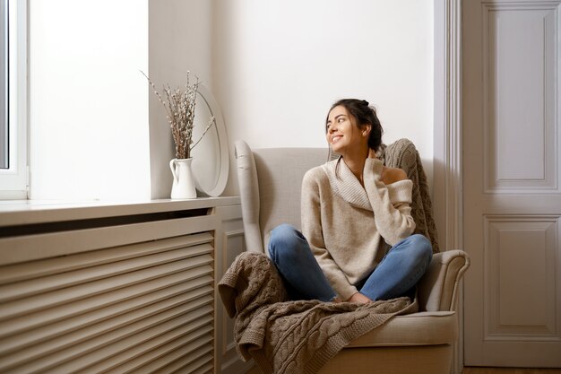 Dame souriante en vêtements à la mode intelligente est assise sur un fauteuil. Souriant, assis dans une atmosphère relaxante à l'intérieur