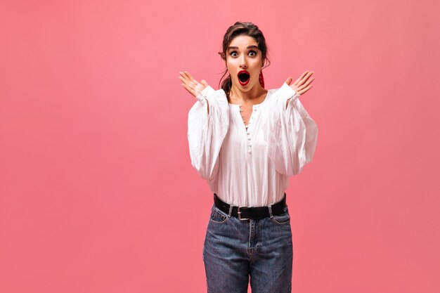 Dame surprise en jeans et chemisier blanc se penche sur la caméra. Fille aux lèvres rouges et dans des vêtements élégants posant sur fond isolé.