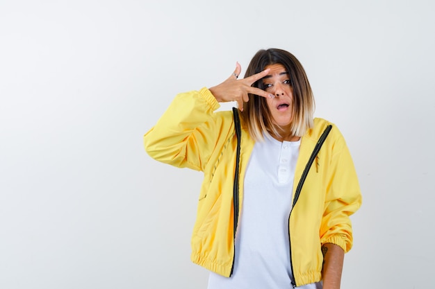 Dame en t-shirt, veste montrant le signe V près de l'œil et regardant perplexe, vue de face.