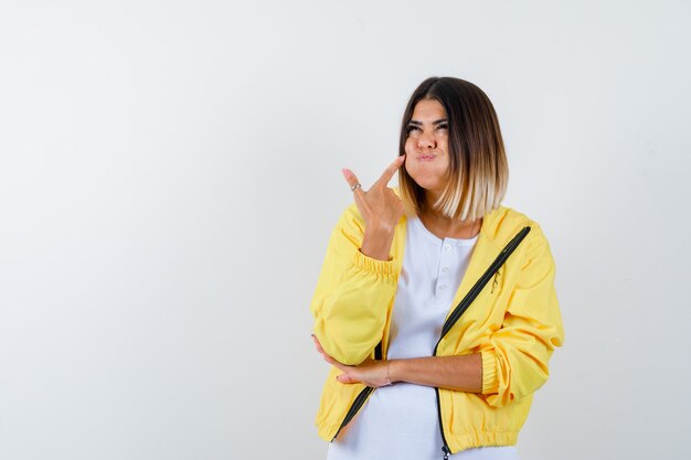 Dame en t-shirt, veste pointant sur sa joue soufflée et à la drôle, vue de face.