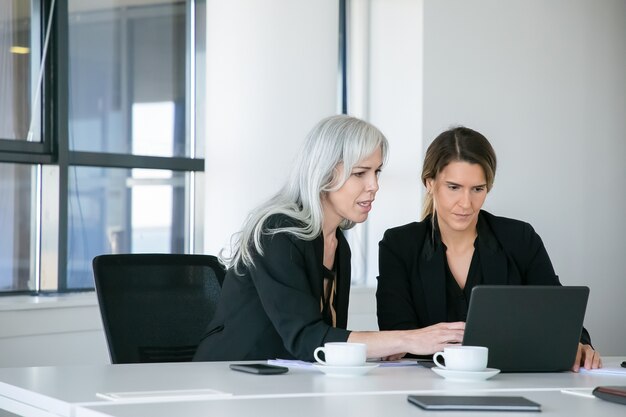 Dames d'affaires ciblées regardant le contenu sur un ordinateur portable assis à table avec des tasses de café et de parler. Concept de travail d'équipe et de communication