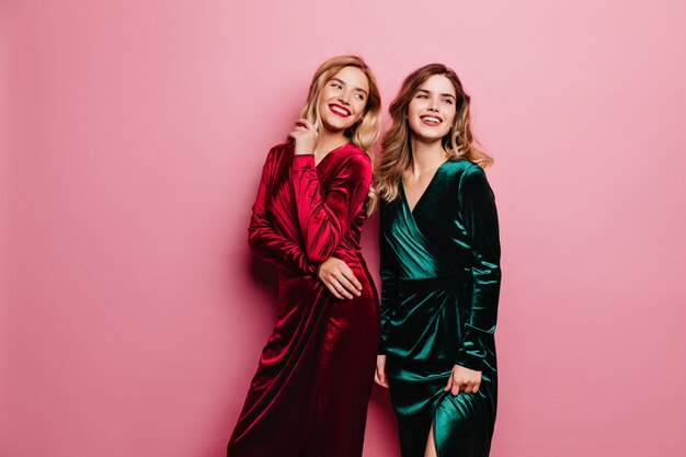 Dames de rêve dans des robes de velours élégantes posant avec un joli sourire. Photo intérieure de jolies filles blanches isolées sur un mur rose.