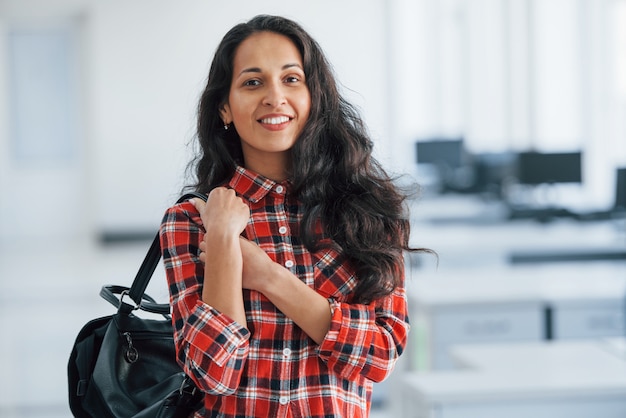 Dans des vêtements décontractés. Portrait de jeune femme séduisante debout au bureau avec sac noir