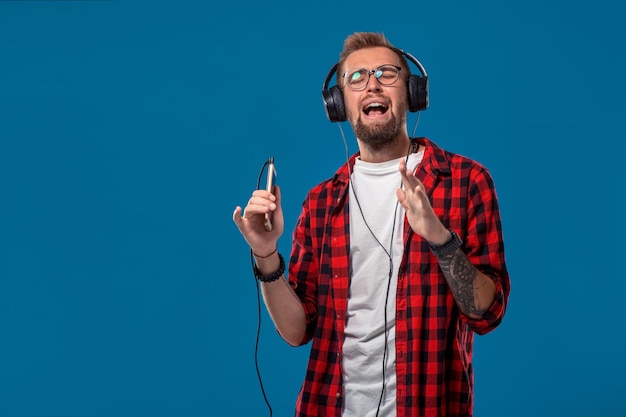 Danse Beau jeune homme élégant dans des écouteurs tenant un lecteur MP3 et dansant en se tenant debout sur fond bleu. Concept émotionnel et humain : jeune homme barbu en chemise à carreaux. Style hipster.