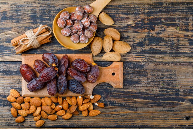 Dates sur une planche à découper avec des amandes pelées et non pelées, des noix dans une cuillère en bois, des bâtons de cannelle vue de dessus sur un fond en bois