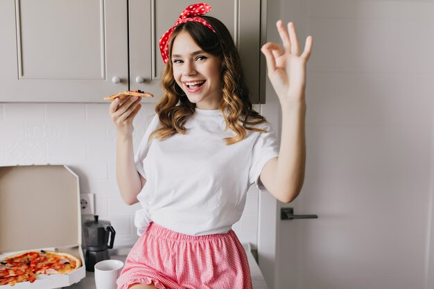 Debonair dame en pyjama mignon appréciant le petit déjeuner. Charmant modèle féminin mangeant de la pizza avec plaisir.