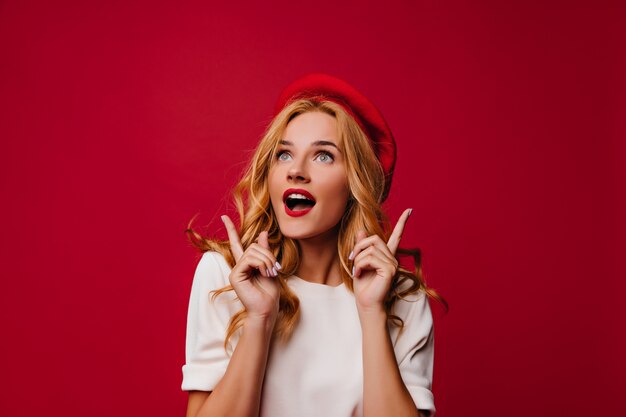 Debonair fille française posant sur un mur rouge. Fille rêveuse en béret élégant exprimant des émotions sincères.