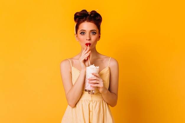 Debonair jeune femme buvant du milkshake. Belle fille rousse en tenue de pin-up debout sur un espace jaune.