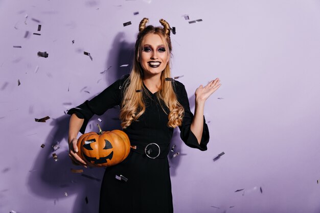 Debonair jeune femme avec un maquillage effrayant se détendre dans le carnaval d'halloween. Rire fille aux cheveux longs en tenue de vampire posant à la fête avec de la citrouille.