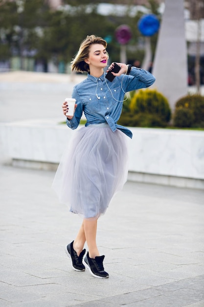 Debout sur la pointe des pieds danseuse dans la rue, vêtue d'une chemise en jean bleu et d'une belle jupe en tulle gris avec des lèvres rose vif et écoutant de la musique.