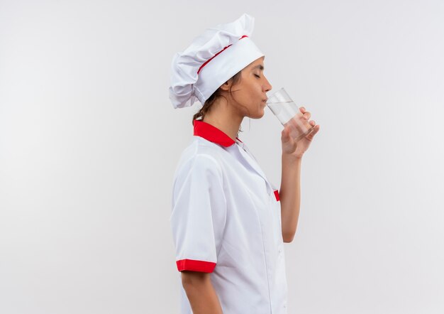 Debout en vue de profil jeune femme cuisinier portant l'uniforme de chef buvait de l'eau en verre avec copie espace