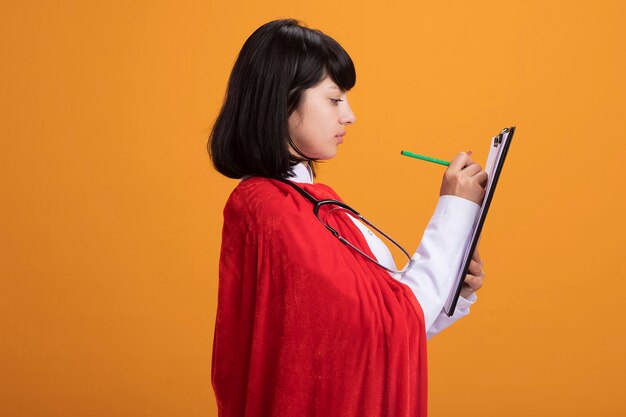 Debout en vue de profil jeune fille de super-héros portant un stéthoscope avec une robe médicale et une cape d'écrire quelque chose sur le presse-papiers