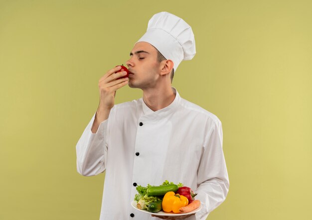 Debout en vue de profil jeune homme cuisinier portant un uniforme de chef tenant des légumes sur une assiette et reniflant la tomate