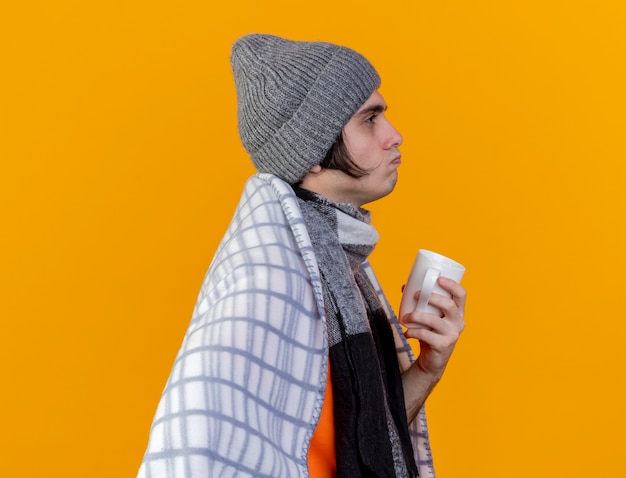 Debout en vue de profil jeune homme malade portant un chapeau d'hiver avec écharpe enveloppé dans un plaid tenant une tasse de thé ¡