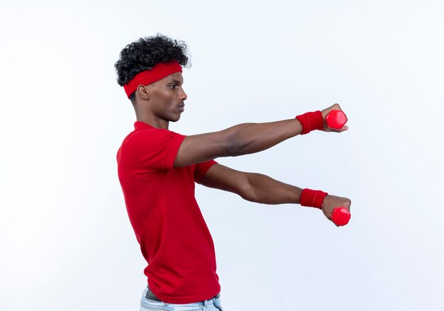 Debout en vue de profil jeune homme sportif afro-américain portant bandeau et bracelet tenant des haltères à côté isolé sur blanc