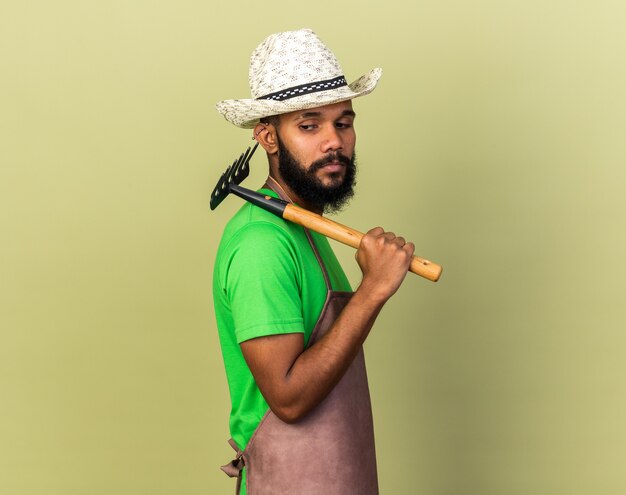 Debout en vue de profil jeune jardinier afro-américain portant un chapeau de jardinage tenant un râteau sur l'épaule isolé sur un mur vert olive