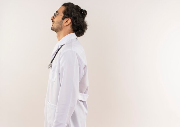 Debout en vue de profil jeune médecin de sexe masculin avec des lunettes optiques portant une robe blanche avec stéthoscope sur mur blanc isolé avec espace copie