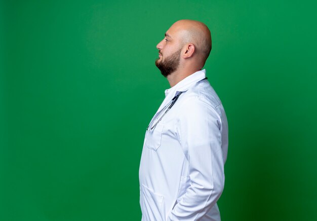 Debout en vue de profil jeune médecin de sexe masculin portant une robe médicale et un stéthoscope isolé sur fond vert avec espace copie