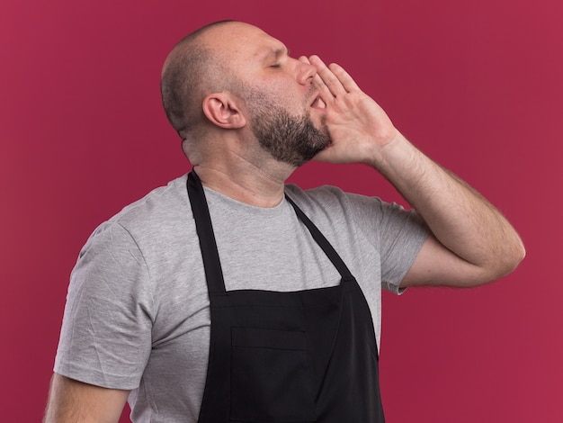 Debout en vue de profil avec les yeux fermés barbier mâle d'âge moyen slave en uniforme appelant quelqu'un isolé sur mur rose