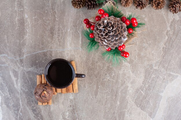 Décorations de fête avec des pommes de pin à côté d'une tasse de thé sur des bâtons de cannelle et un biscuit sur du marbre.