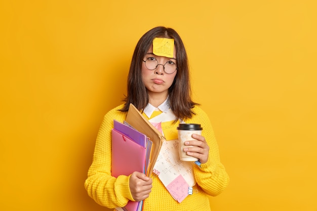 Déçu une femme diplômée fatiguée se prépare pour la session d'examen a la date limite entourée de notes autocollantes et de papiers tient une tasse de café jetable a mécontent mécontent expression se tient à l'intérieur