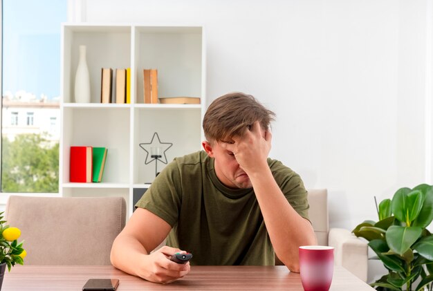 Déçu jeune bel homme blond est assis à table avec téléphone et tasse