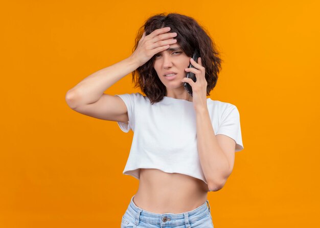 Déçu jeune belle femme mettant la main sur le front et tenant le téléphone mobile sur un mur orange isolé avec espace de copie