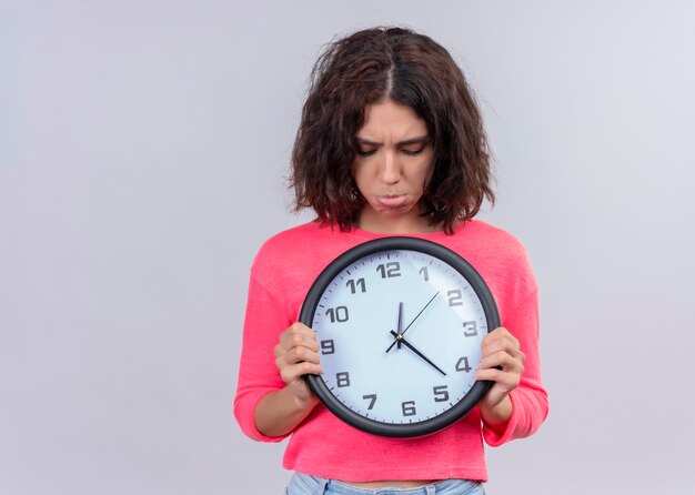 Déçu jeune belle femme tenant horloge et en le regardant sur un mur blanc isolé avec copie espace