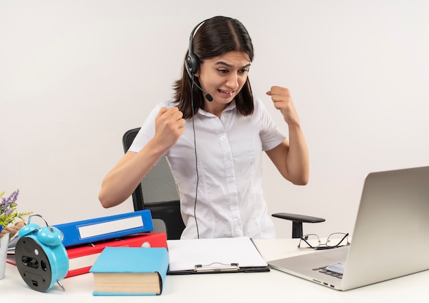 Déçu jeune femme en chemise blanche et un casque avec un microphone serrant les poings assis à la table avec des dossiers et un ordinateur portable sur un mur blanc