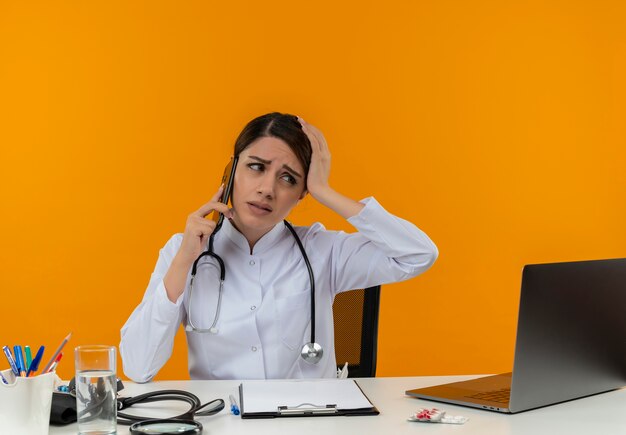 Déçu jeune femme médecin portant une robe médicale et un stéthoscope assis au bureau avec des outils médicaux et un ordinateur portable parlant au téléphone à côté isolé sur mur jaune