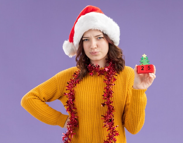 Déçu jeune fille slave avec bonnet de Noel et avec guirlande autour du cou tenant ornement de sapin de Noël isolé sur fond violet avec espace copie