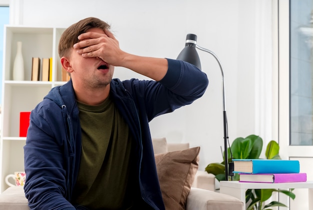 Photo gratuite déçu jeune homme beau blond est assis sur un fauteuil mettant la main sur le visage fermant les yeux à l'intérieur du salon